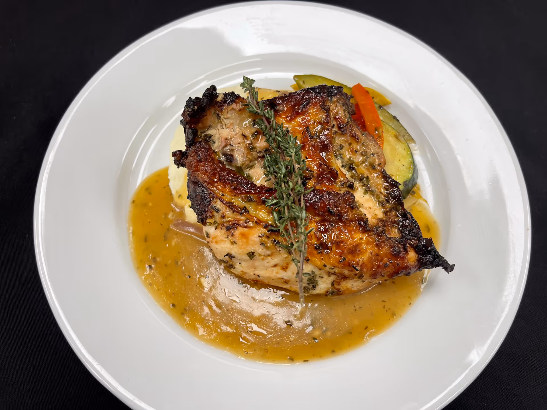 A white plate with a serving of roasted chicken breast garnished with a sprig of thyme, accompanied by mashed potatoes, sautéed vegetables including zucchini and carrots, and covered with a light brown gravy sauce, all set against a black tablecloth background.