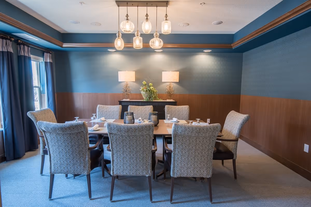 A dining room with a rectangular wooden table surrounded by eight upholstered chairs. The table is set with placemats, napkins, and glassware. Two table lamps and a floral arrangement are on a sideboard against the wall. The walls are painted blue with a wooden wainscoting, and there are large windows with blue curtains on the left side. Several pendant lights hang from the ceiling above the table.