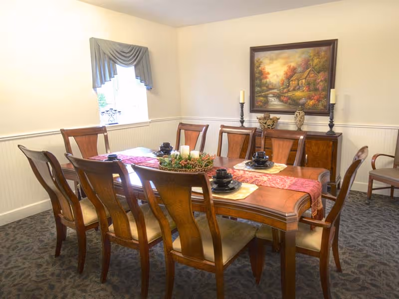 A dining room with a wooden dining table set for six, featuring black cups and saucers, a red table runner, and a centerpiece with candles and greenery. The room has beige walls, a window with a blue valance, a sideboard with decorative items, and a framed painting of a cottage and autumn trees on the wall.