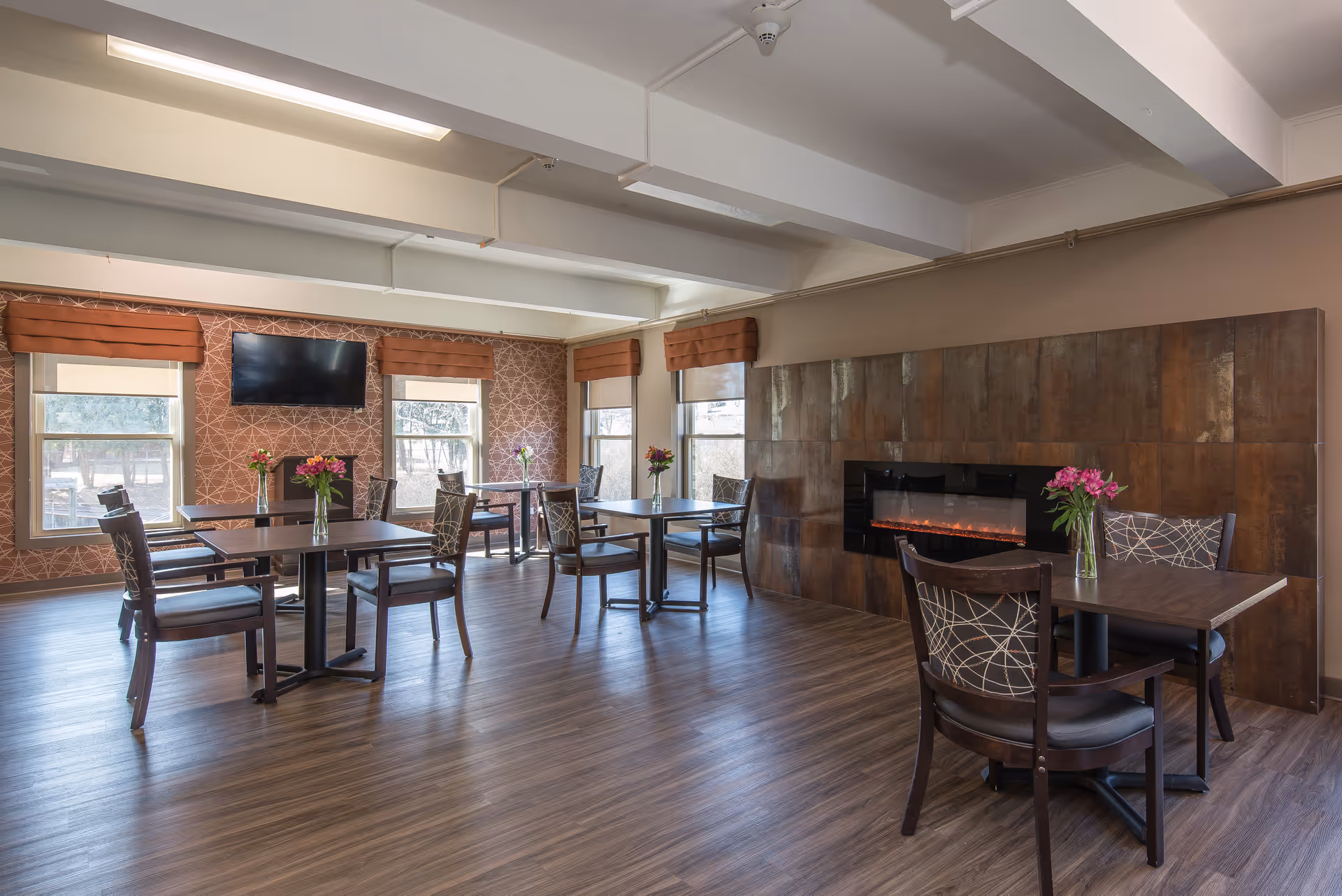 A dining room with several tables and chairs arranged neatly. Each table has a vase with flowers. The room features large windows with brown valances, a wall-mounted TV, and a modern electric fireplace set into a tiled wall. The floor is wood, and the walls have decorative patterns.