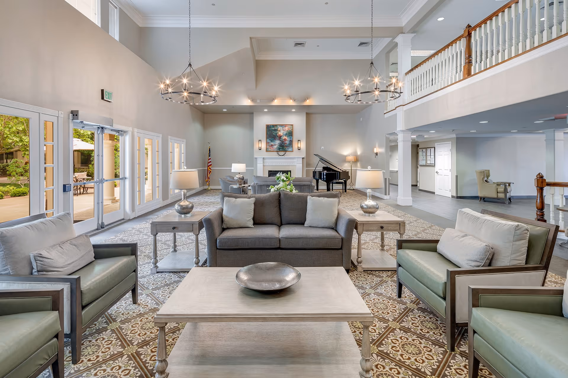 Spacious and well-lit living room area with comfortable seating including a gray sofa and green armchairs arranged around a wooden coffee table. The room features large windows and glass doors letting in natural light, two modern chandeliers hanging from a high ceiling, a fireplace with artwork above it, and a grand piano in the background. The floor has a patterned rug and there is a balcony with white railings overlooking the space.