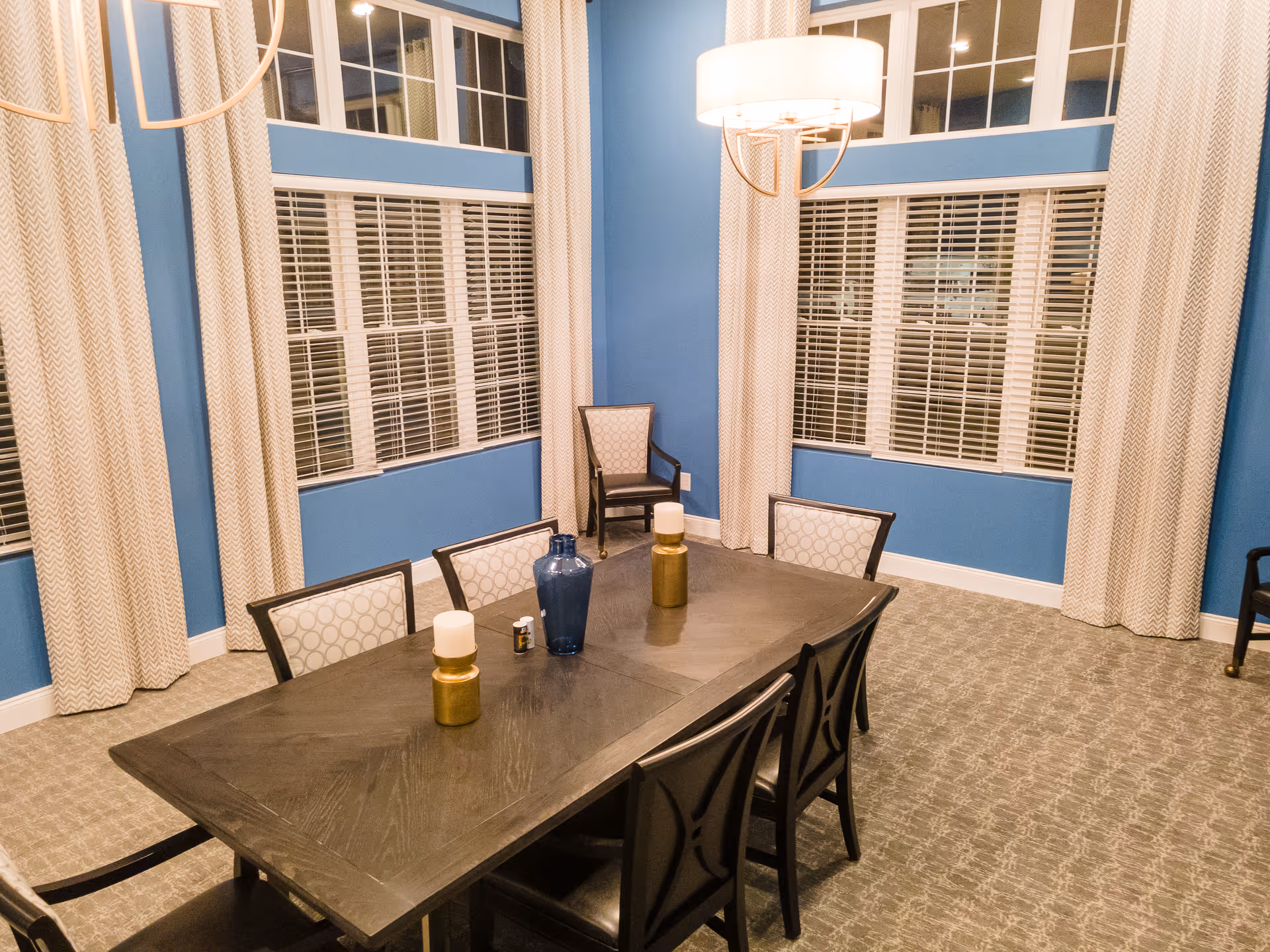 Dining room with a dark wooden table and chairs, blue walls, large windows with blinds and curtains, and overhead lighting.