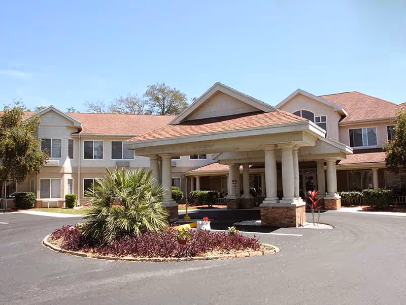 Exterior view of The Springs at Lady Lake Assisted Living facility showing a two-story building with a covered entrance supported by columns, surrounded by landscaped plants and a circular driveway under a clear blue sky.