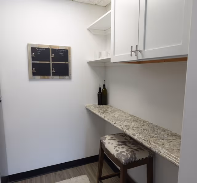 Small kitchen nook with granite counter, white cabinets, shelves, and a stool.
