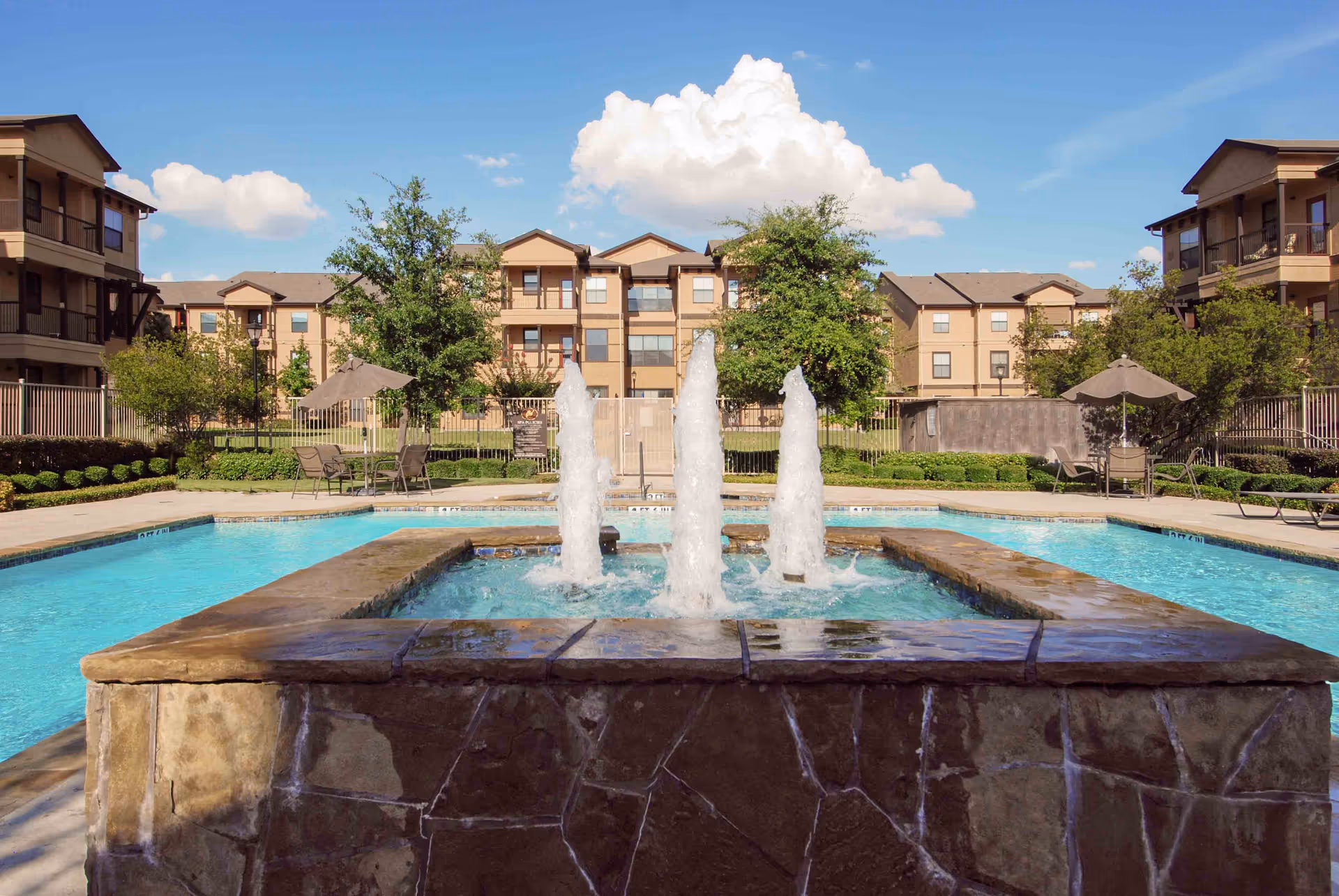 Outdoor swimming pool with a central stone fountain, lounge chairs and umbrellas, and apartment buildings in the background under a blue sky.