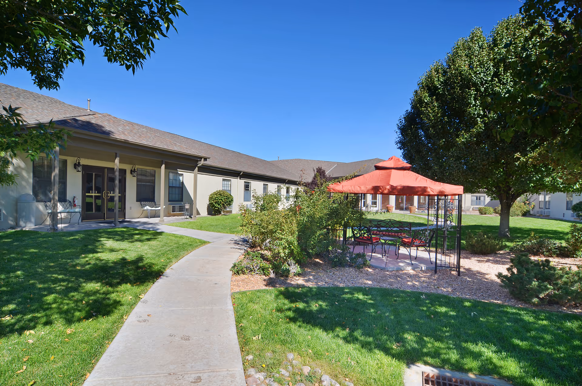 Sunny courtyard with a paved walkway, lawn, a red-canopied seating gazebo and surrounding single-story building.