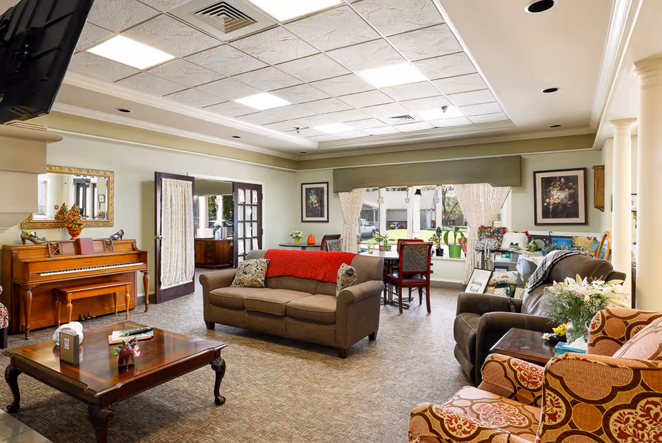 A cozy living room area in an assisted living facility featuring a brown sofa with a red throw, a wooden coffee table, a piano with a bench, and several armchairs. The room has large windows with curtains, framed artwork on the walls, and a dining table with chairs near the window. The space is well-lit with ceiling lights and has a warm, inviting atmosphere.