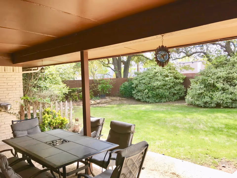 Covered patio area with a rectangular outdoor table and six cushioned chairs, overlooking a green lawn with bushes and trees in the background.