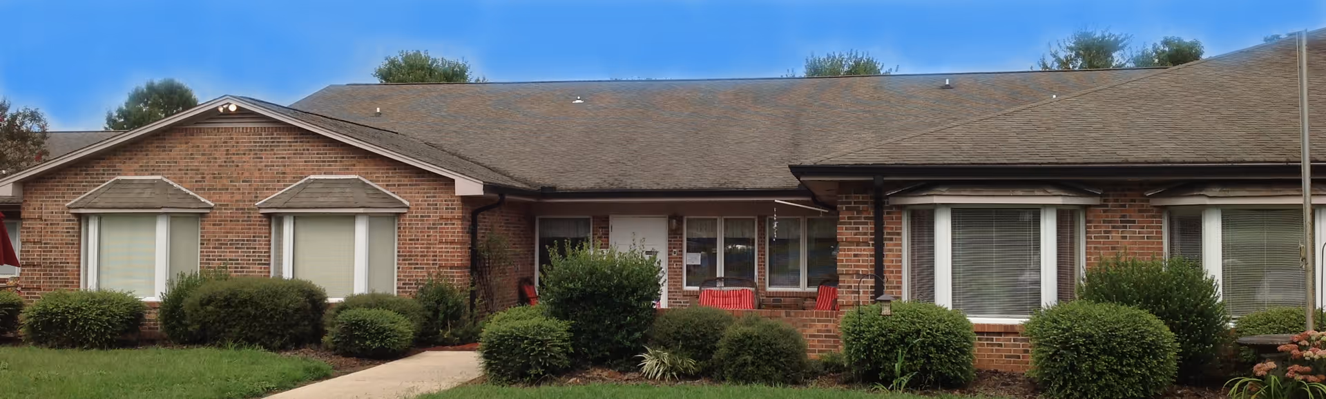 Front exterior of a single-story brick senior living building with a small covered entrance, windows, shrubs, and a walkway.