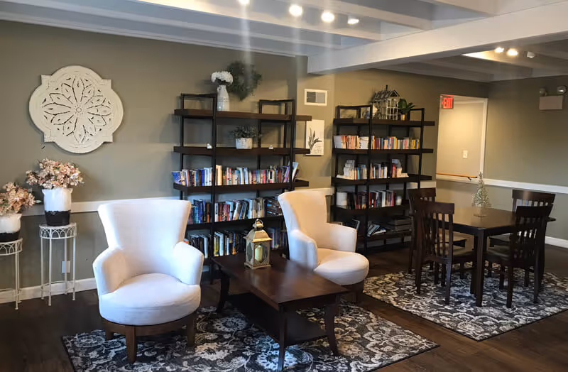 A cozy interior common area with two white upholstered armchairs around a wooden coffee table, two dark wooden bookshelves filled with books against the wall, a dark wooden dining table with four chairs on a patterned rug, and decorative plants and wall art in the background.
