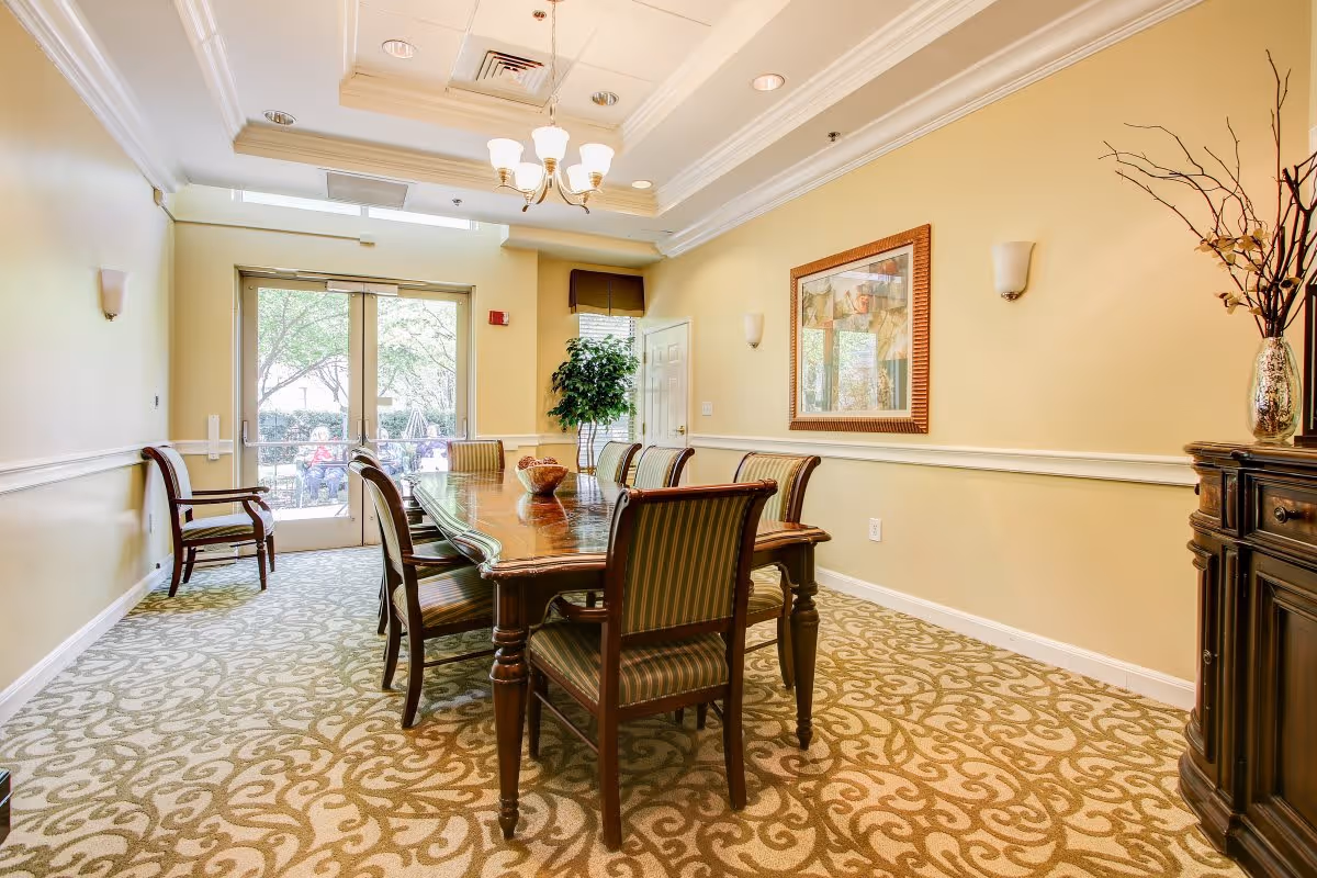 A well-lit dining room with a long wooden table surrounded by eight striped upholstered chairs. The room has beige walls with white trim, a patterned carpet, a chandelier hanging from a tray ceiling, a framed artwork on the wall, a potted plant near a window, and glass double doors leading outside.