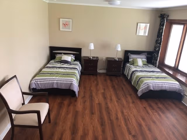 A bedroom with two single beds featuring matching green, gray, and white striped bedding. Each bed has a pillow and a small cushion. Between the beds are two dark wooden nightstands, each with a white lamp. There is a wooden chair with a beige cushion on the left side of the room. The room has wooden flooring, beige walls, two framed pictures on the wall, and a large window with dark curtains on the right side.