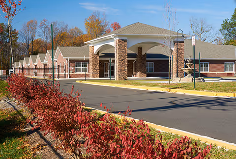 Exterior view of a single-story assisted living facility with a covered entrance supported by stone pillars. The building is surrounded by a paved driveway and landscaped with red-leaved bushes and trees with autumn foliage under a clear blue sky.