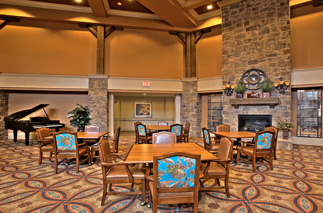 Spacious common area with multiple wooden tables and chairs featuring blue floral upholstery, a grand piano in the corner, stone pillars, a large stone fireplace with decorative items on the mantel, and patterned carpet flooring under warm lighting.