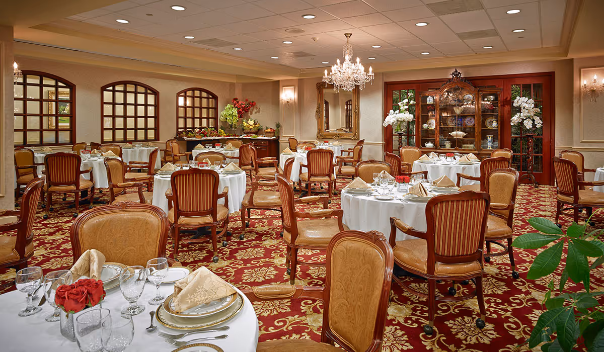Elegant formal dining room with round tables set with white linens, place settings, and wooden chairs on a patterned red carpet.