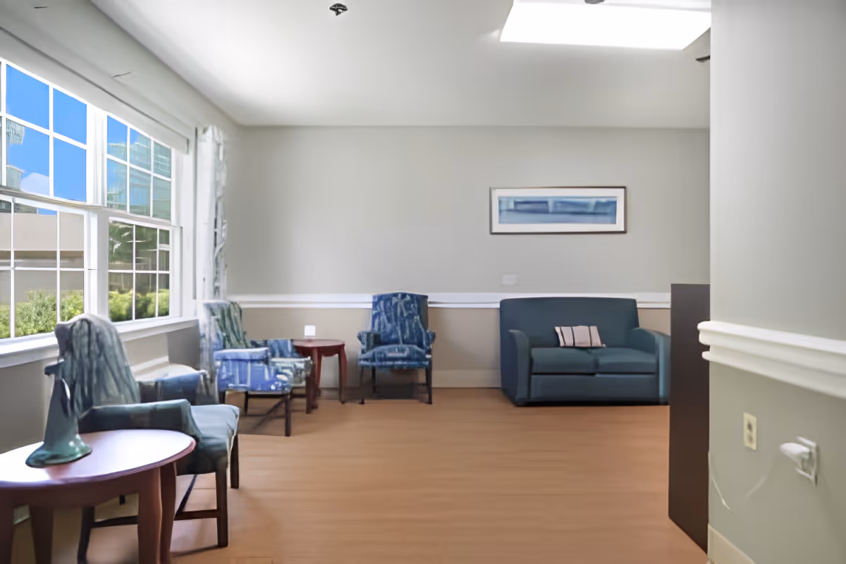 A bright sitting area with large windows on the left letting in natural light. The room has several upholstered chairs in blue tones, a small blue loveseat with a pillow, and two small wooden side tables. A framed abstract artwork hangs on the wall above the loveseat. The floor is wooden, and the walls are painted light gray with white trim.