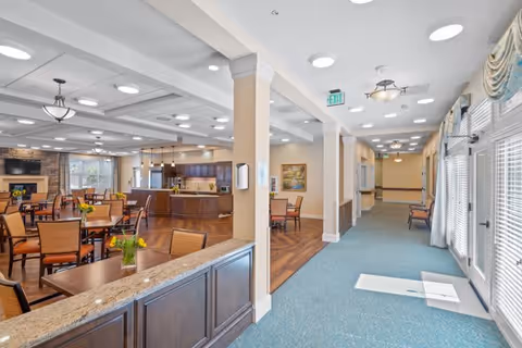 Interior view of a senior living facility showing a spacious common area with multiple tables and chairs arranged for dining or socializing. The area features a kitchen or serving counter with granite countertops, a fireplace in the background, and large windows allowing natural light. Adjacent to the dining area is a carpeted hallway with seating along the walls and glass doors leading outside.
