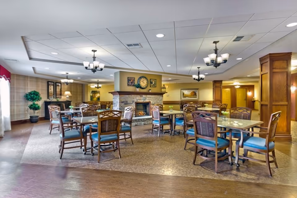 Spacious communal dining area with multiple tables and chairs arranged around a central stone fireplace under chandelier lighting.