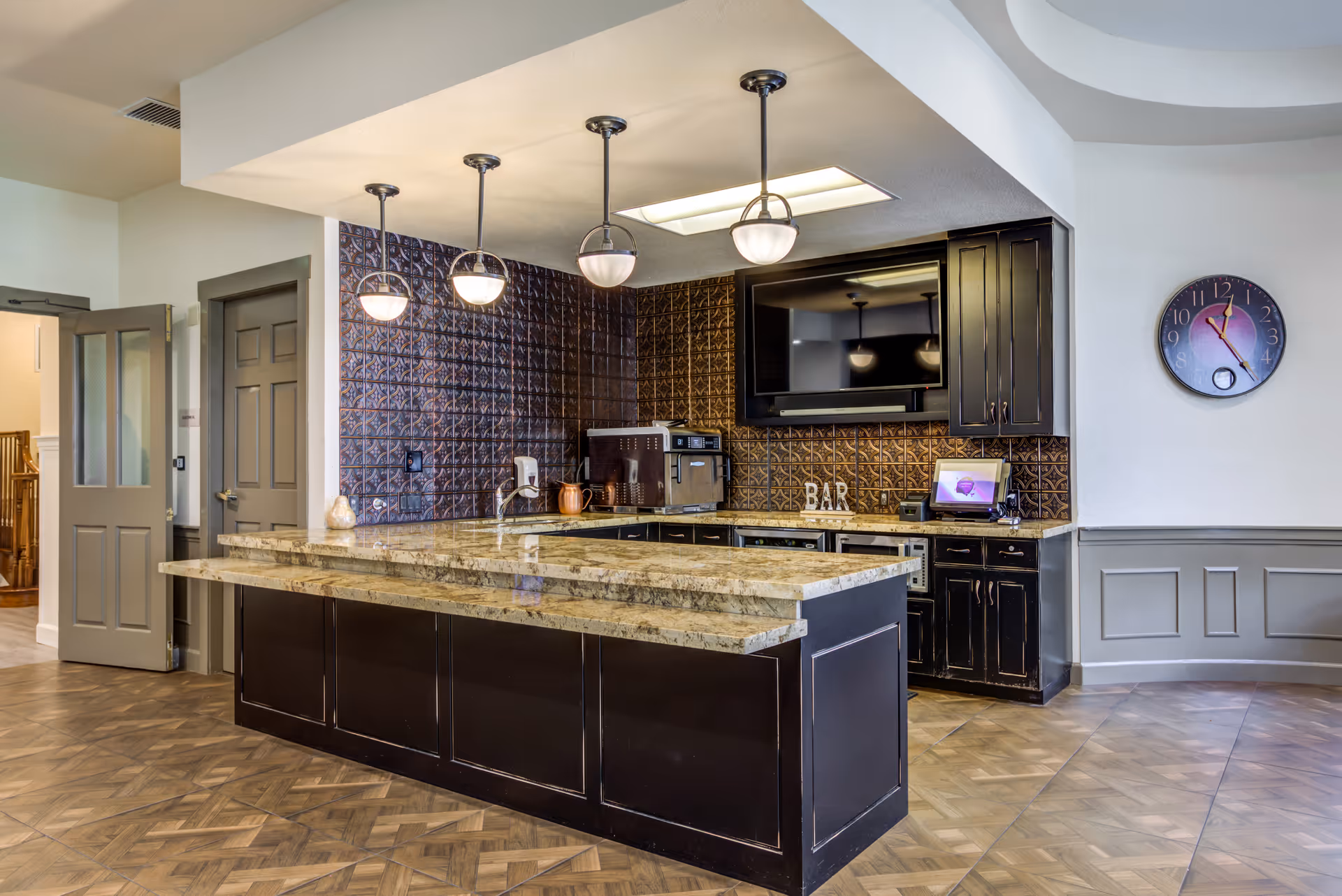Modern kitchen area with a large marble countertop island, dark cabinetry, three pendant lights hanging from the ceiling, a wall-mounted TV, and a clock on the wall showing 12:25. The floor has a patterned wood design and there is a door open to another room.