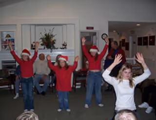 A group of people in a common room, with some wearing red shirts and Santa hats, raising their arms in a celebratory or dance pose. Several seated individuals watch the activity. The room has white walls, framed pictures, and a flower arrangement on a shelf.
