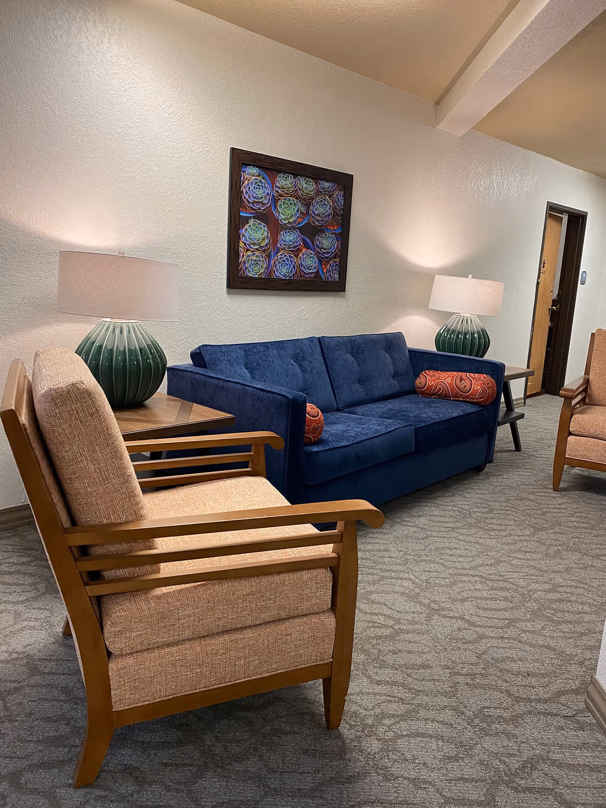 A cozy seating area in a senior living facility featuring a blue upholstered sofa with two red patterned bolster pillows, two beige cushioned armchairs with wooden frames, two green ceramic table lamps with white shades on wooden side tables, and a colorful framed artwork of succulents on a white textured wall.