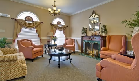 Sunlit lounge with upholstered armchairs arranged around a round coffee table in front of a decorative fireplace and arched windows.