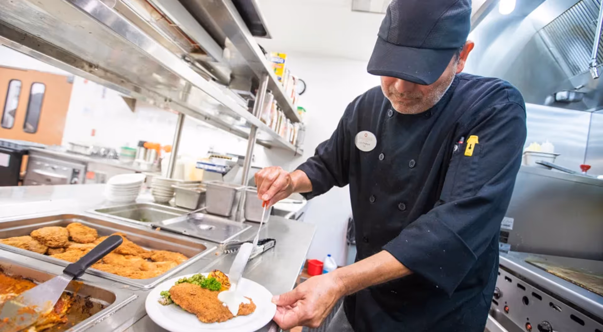 A chef in a commercial kitchen plating a breaded cutlet with sauce from a hot food counter.