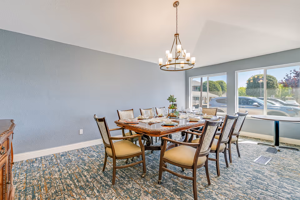 A dining room with a large wooden table set for eight people with plates, glasses, and a centerpiece. The room has a blue-gray wall, patterned carpet, large windows showing parked cars outside, and a chandelier hanging above the table.
