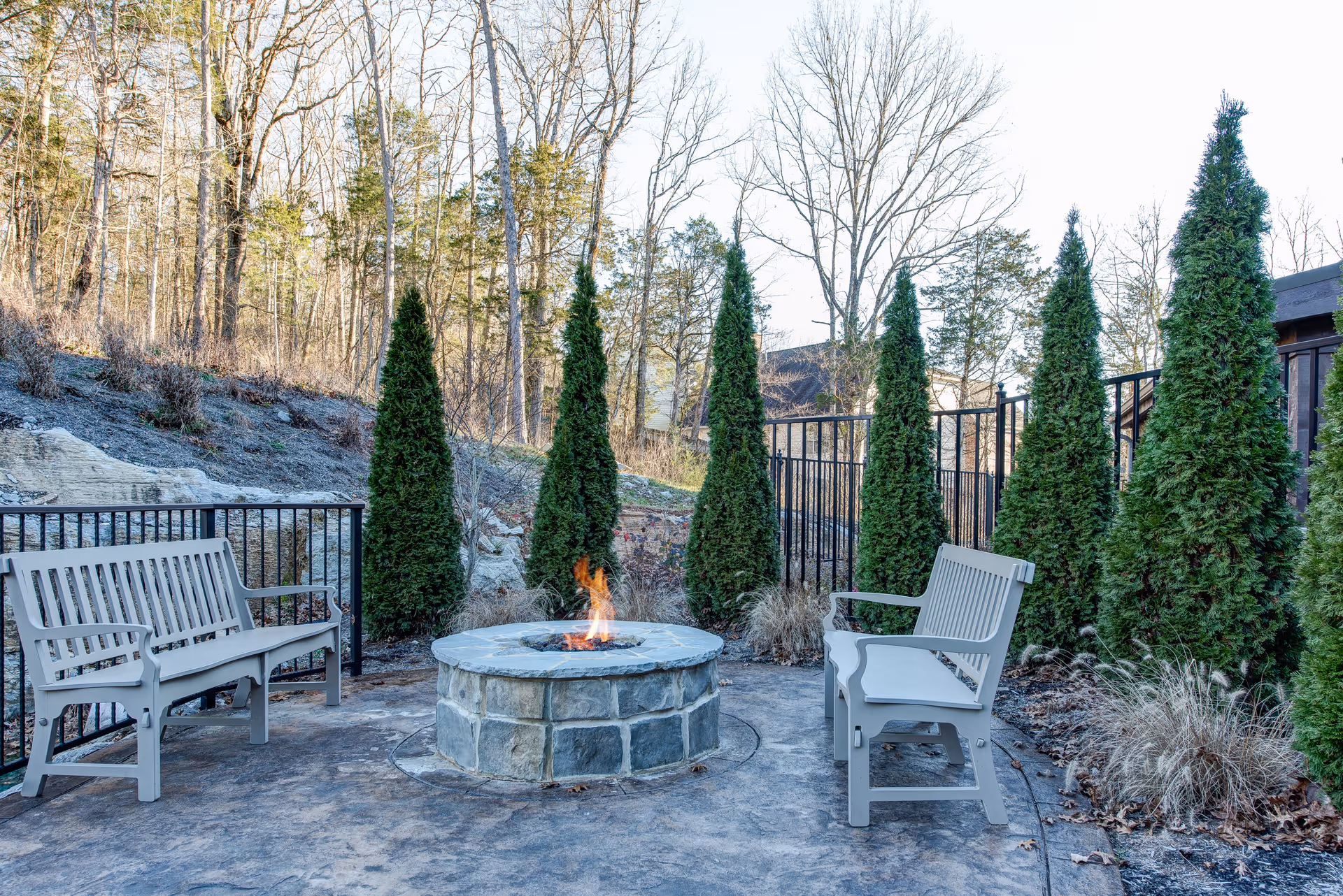 Outdoor seating area with two gray benches facing a stone fire pit with a small flame, surrounded by tall evergreen trees and a black metal fence, with leafless trees and a hillside in the background.