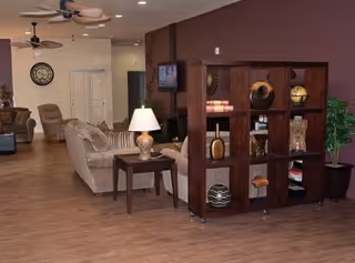 A spacious living room area with wooden flooring, beige sofas, a side table with a lamp, a large wooden shelving unit with decorative items, a wall-mounted TV, ceiling fans, and a clock on the far wall.