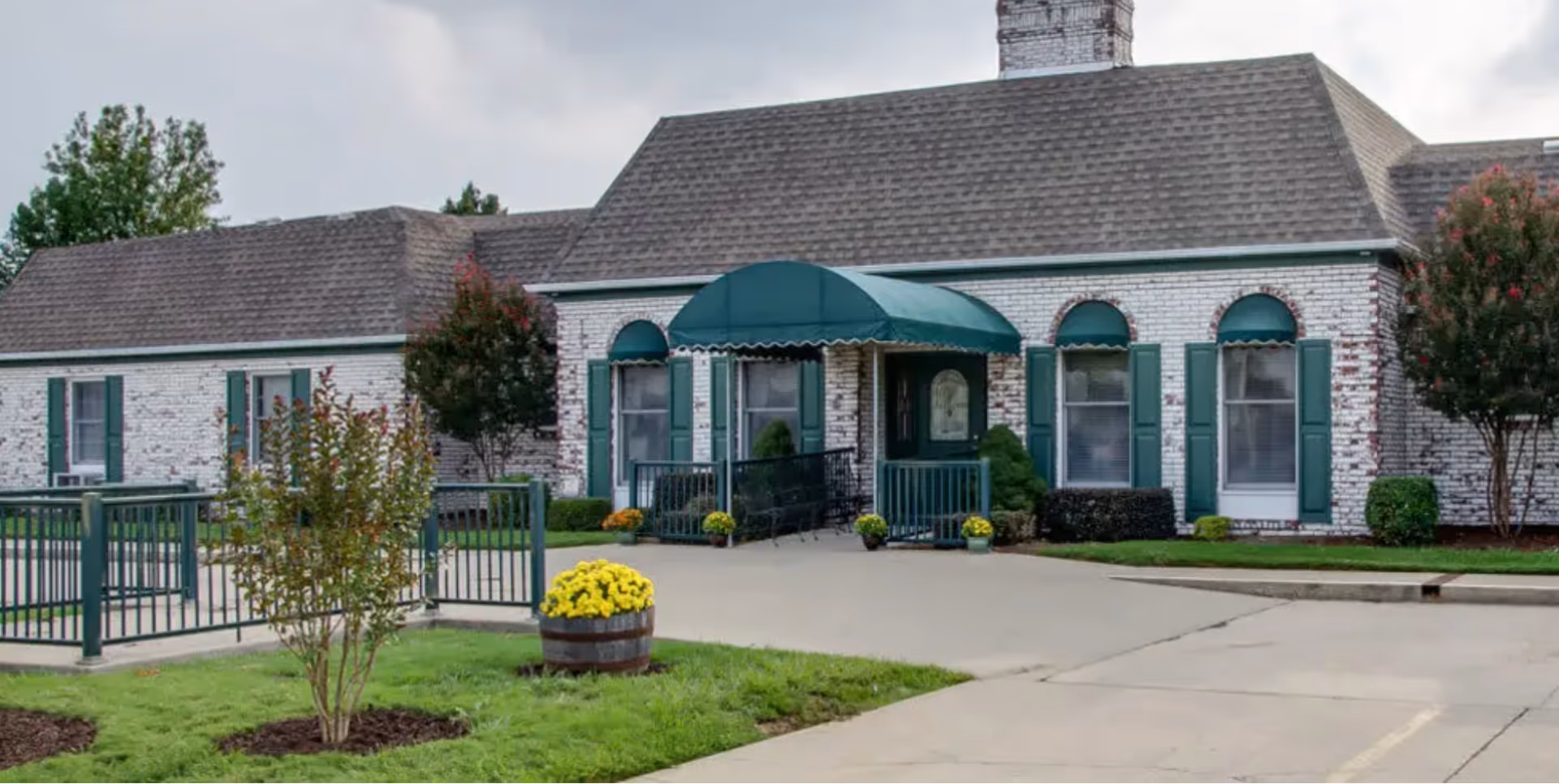 Exterior view of a single-story senior living facility building with white brick walls and green shutters. The entrance has a green awning and a ramp with railings. There are small trees, shrubs, and flower pots with yellow flowers near the entrance and along the driveway.