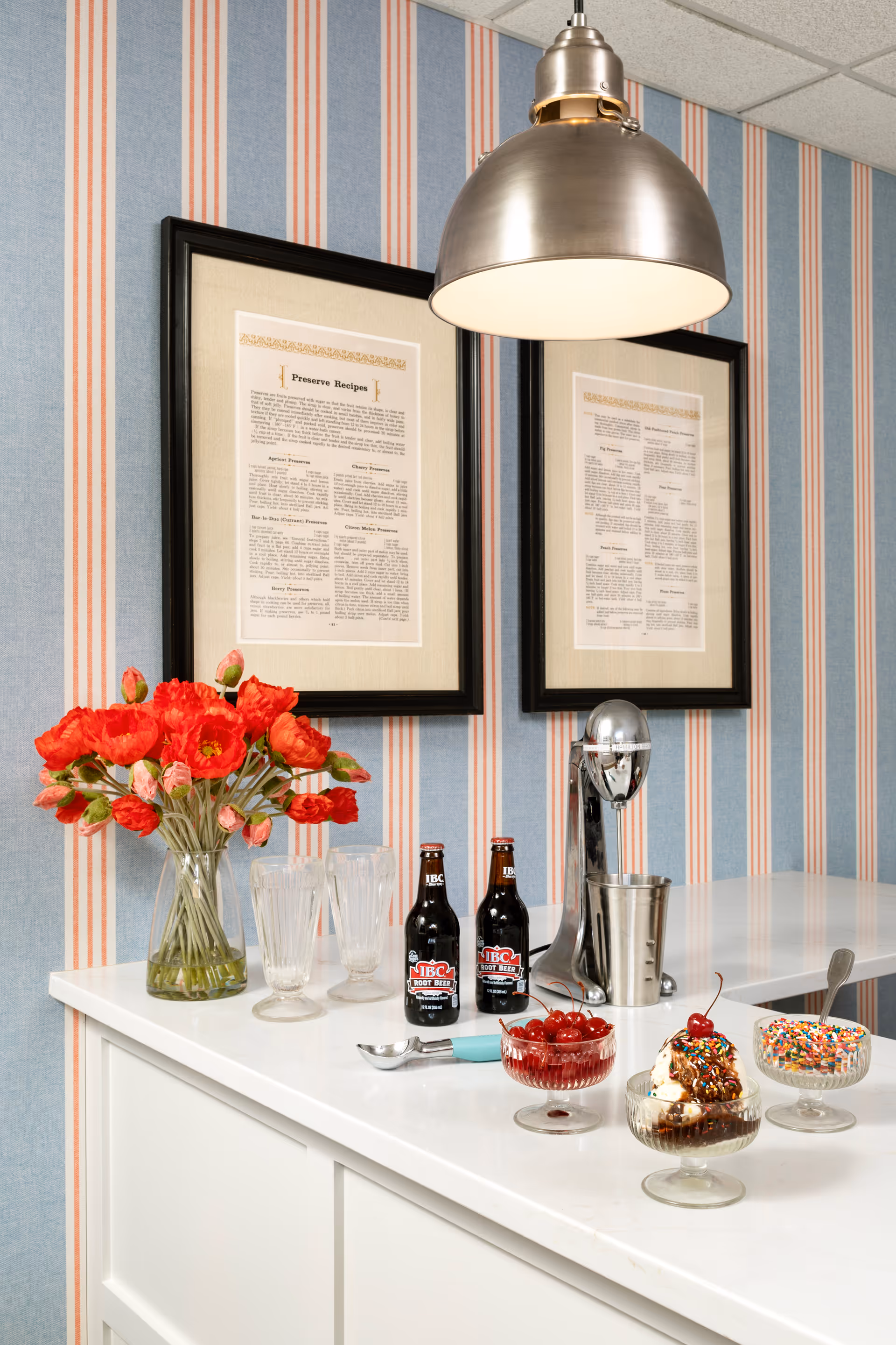 A countertop with ice cream sundaes, bottles of soda, a vase of red flowers, a soda dispenser and framed prints on striped wallpaper beneath a pendant light.