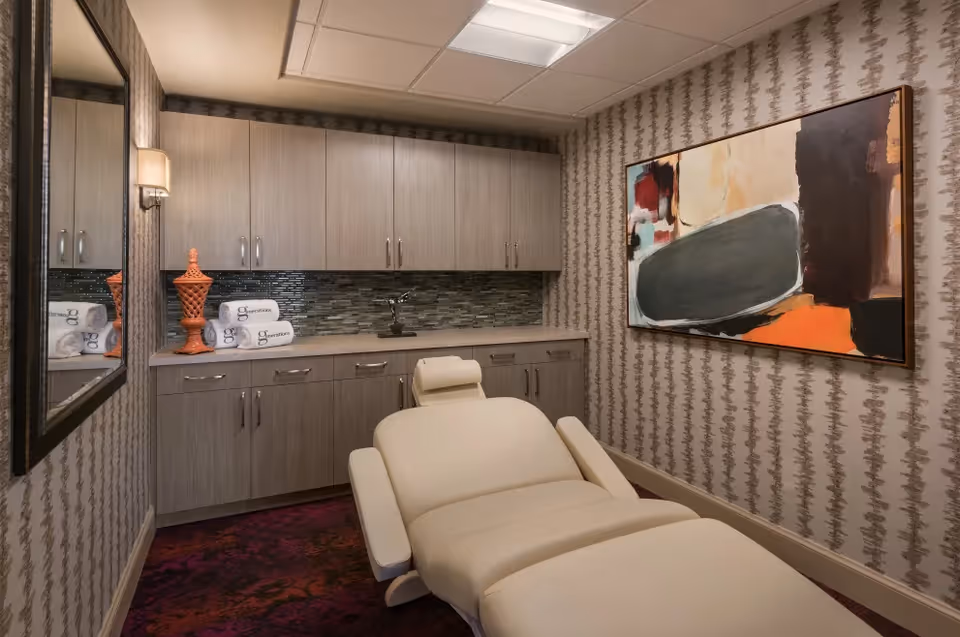 A small spa treatment room with a reclining massage chair, built-in cabinets, decorative artwork and patterned wallpaper.