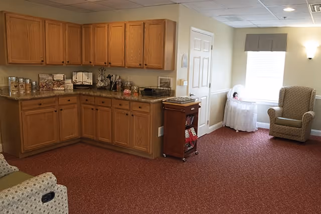 A cozy interior room with wooden cabinets and a countertop on the left side, containing various jars and kitchen items. The room has a red carpeted floor, a window with a valance, a comfortable armchair, and a small table with a doll in a white dress near the window.