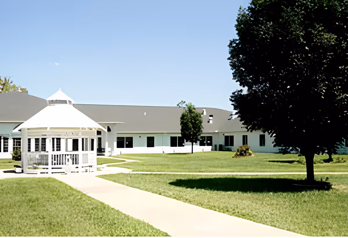 Outdoor view of a senior living facility with a white gazebo on the left, a paved walkway leading towards a single-story building with multiple windows, and a large tree casting a shadow on the right side of the grassy lawn under a clear blue sky.