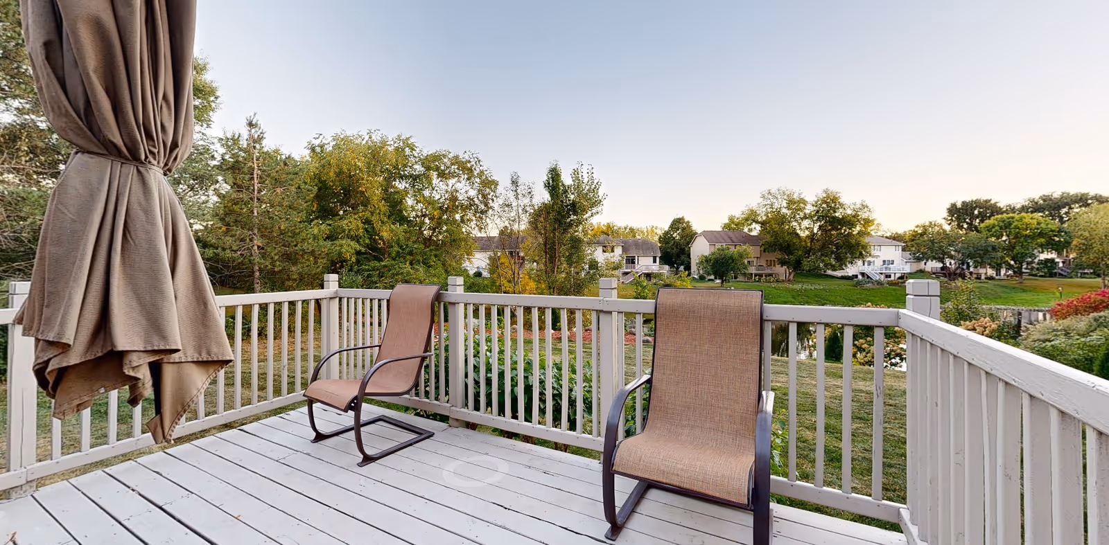 A wooden deck with two outdoor chairs and a closed umbrella overlooking a pond and nearby houses and trees.