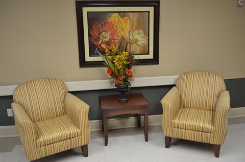 Two striped armchairs with yellow, green, and red tones are positioned on either side of a small wooden table holding a floral arrangement. A framed painting of large flowers in red, yellow, and white hangs on the beige wall behind the chairs.