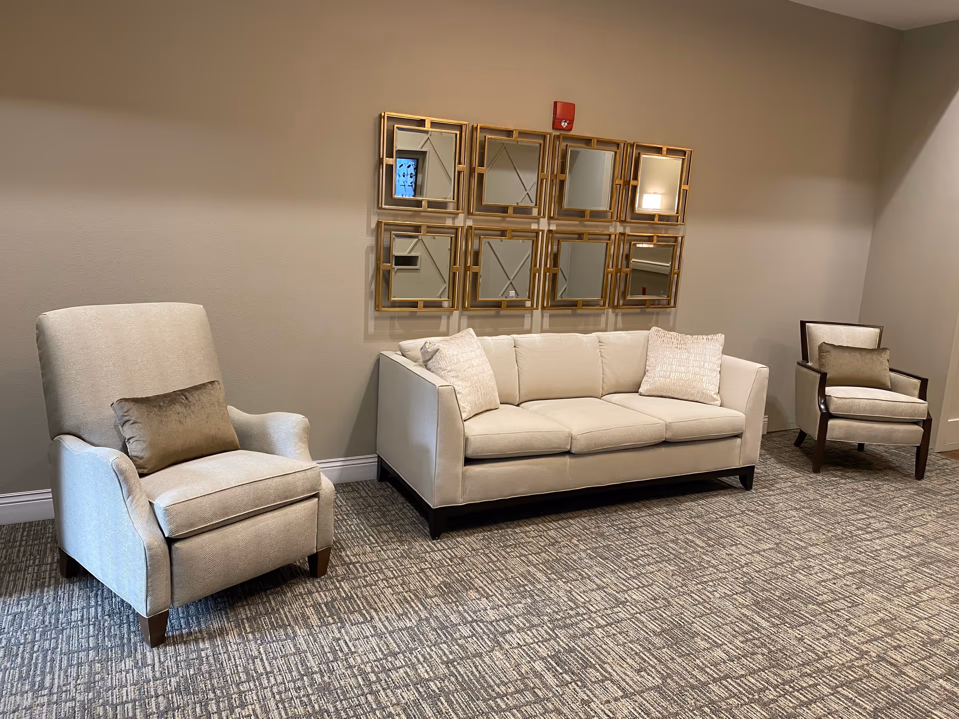 A seating area with a beige sofa and two beige armchairs, each with a decorative pillow. Above the sofa is a wall decoration consisting of nine square mirrors with gold frames arranged in a grid. The room has beige walls and a patterned carpeted floor.