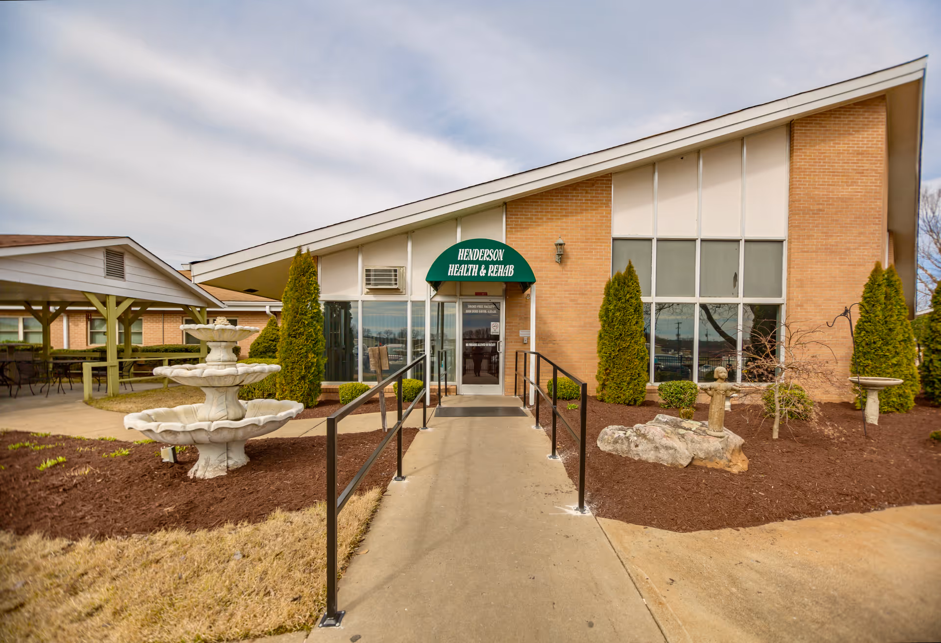 Front entrance of Henderson Health & Rehab center with a green awning sign, access ramp, fountain, and landscaped beds.