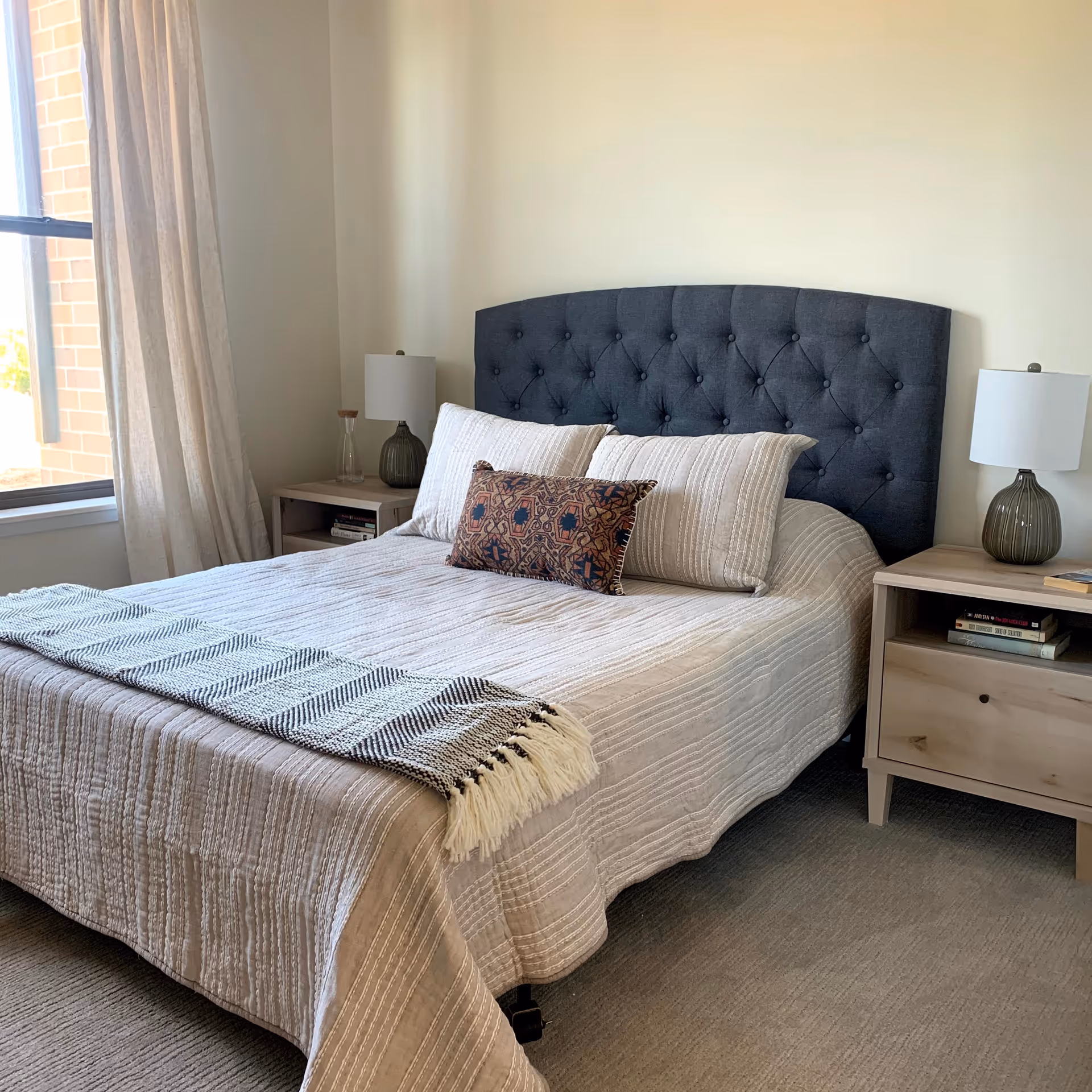 A neatly made bed with a dark tufted headboard, beige striped bedding, and a decorative pillow in front. There are two wooden nightstands on either side of the bed, each with a lamp and some books. A window with light curtains is visible on the left side of the room.