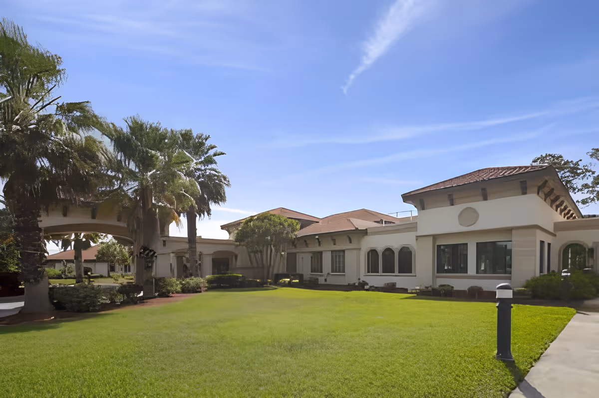 Exterior view of a single-story building with a red-tiled roof, surrounded by green grass and palm trees under a clear blue sky.