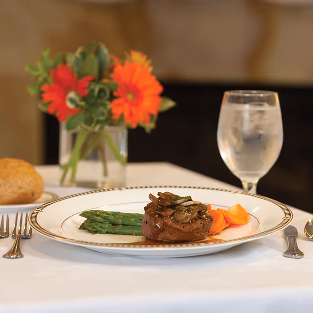 A plated steak topped with mushrooms served with asparagus and carrots on a white tablecloth, beside a glass of water and a vase of orange flowers.