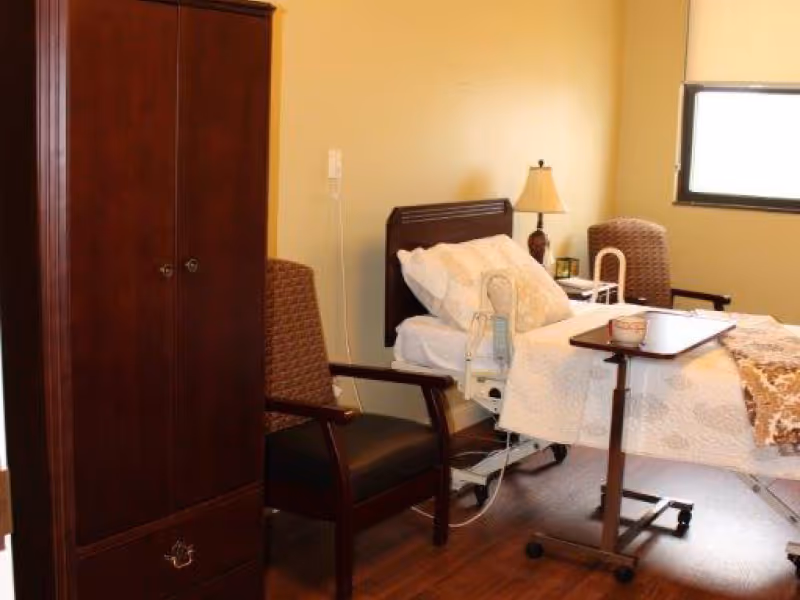 A furnished patient bedroom featuring an adjustable hospital bed with bedding and an overbed table, two armchairs, a wardrobe, and a bedside lamp.