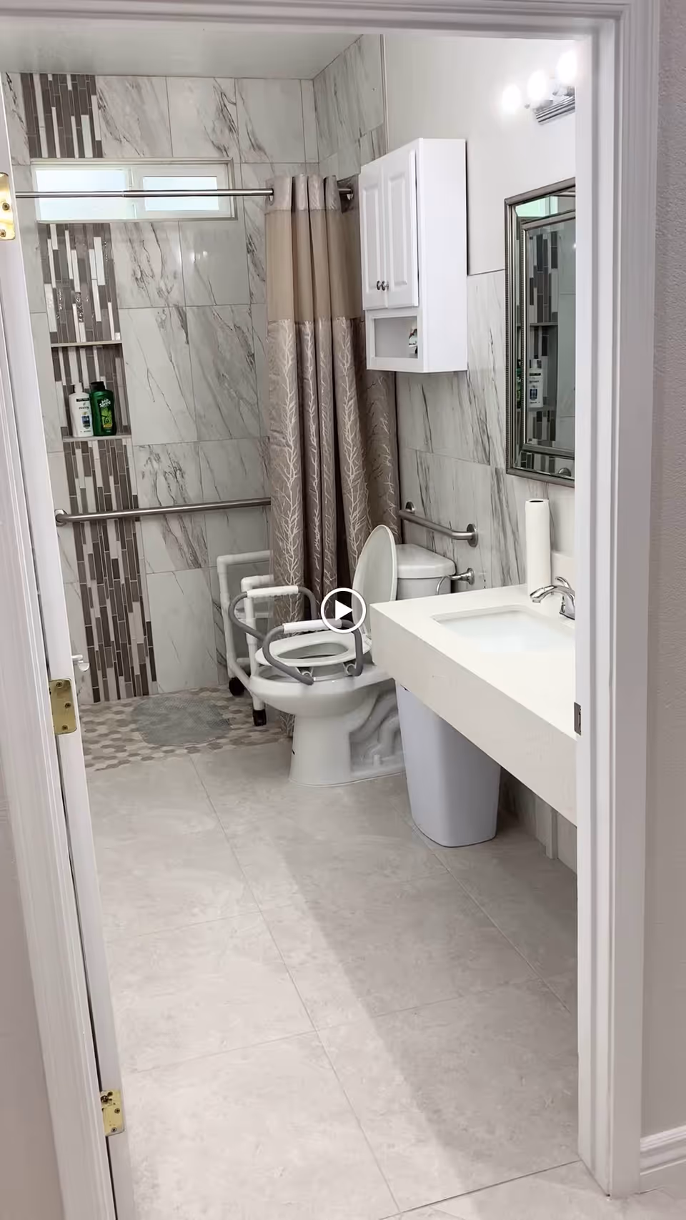 A modern bathroom with light-colored tiled floors and walls. There is a toilet with an attached safety frame, a white sink with a countertop, a wall-mounted cabinet above the toilet, and a mirror with lights above the sink. The shower area has a curtain and a tiled accent wall with grab bars and toiletries.