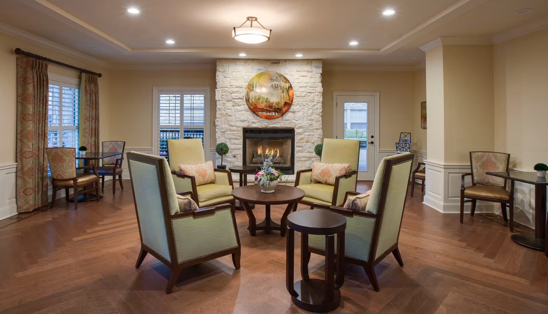 A cozy senior living facility common area with four green upholstered armchairs arranged around a round wooden coffee table with a floral centerpiece. The room features a stone fireplace with a circular decorative piece above it, wooden flooring, and additional seating with small tables near the windows and walls. The space is warmly lit with recessed ceiling lights.