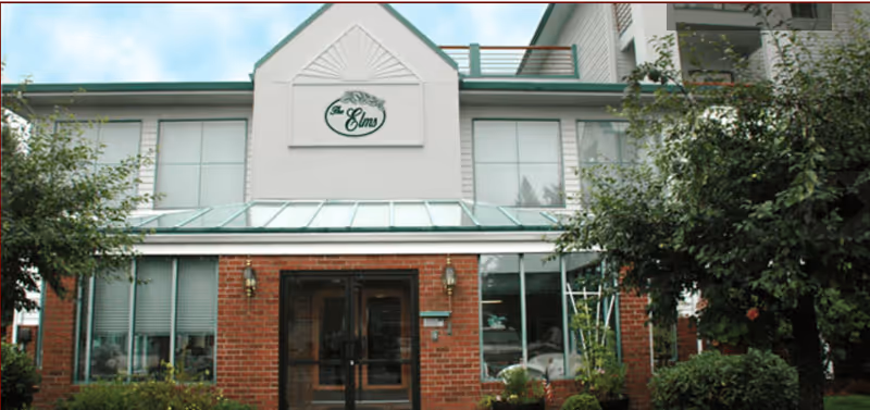 Front exterior view of Elms Residential Center building with brick lower facade, large windows, green metal roof, and trees and shrubs in front.