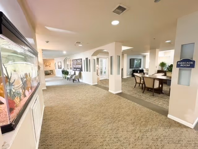 Interior view of a senior living facility hallway with carpeted floors and white walls. On the left side, there is a large aquarium built into the wall. The hallway leads to a seating area with chairs and tables. A sign on the right wall indicates the 'Cascade Room'. The space is well-lit with ceiling lights and has decorative plants and framed pictures on the walls.