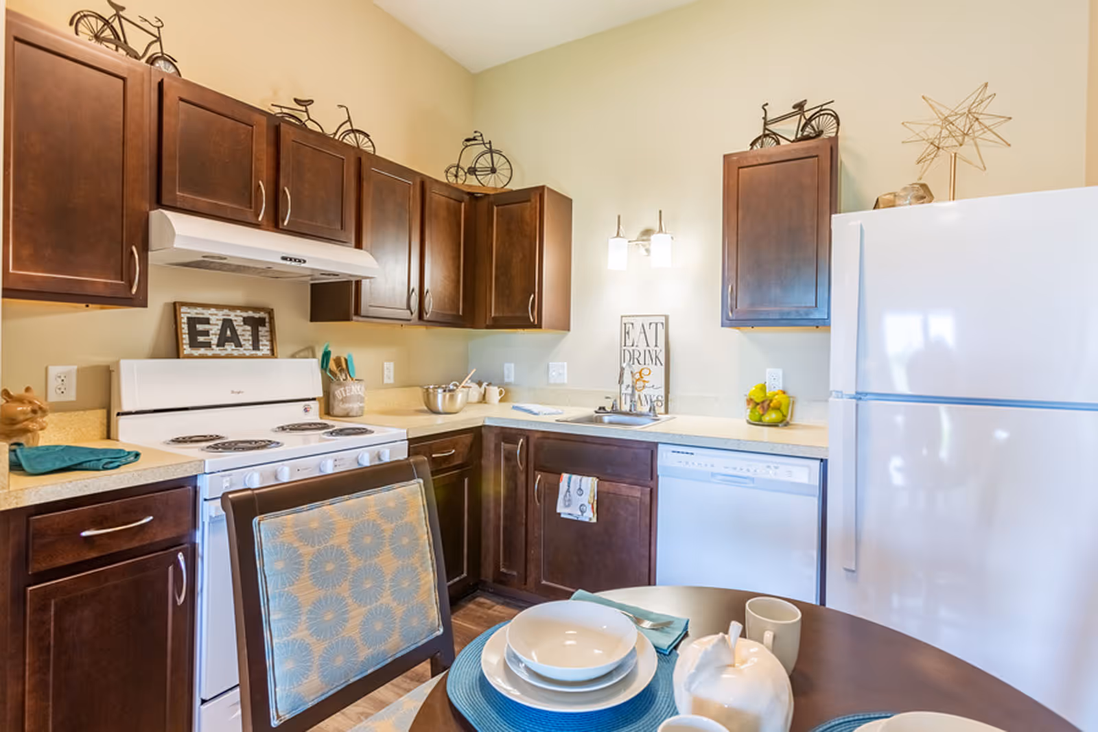 A bright kitchen with dark wood cabinets, a white stove, dishwasher, and refrigerator. The countertop has a bowl, utensils, and a small container of fruit. Decorative metal bicycles and a star sculpture are placed on top of the cabinets. A dining table with blue placemats, white dishes, and a ceramic teapot is in the foreground.