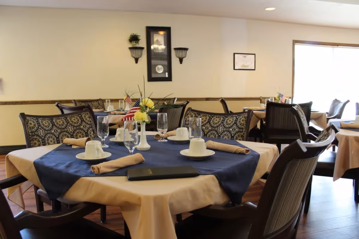 Dining room with tables covered in beige tablecloths and blue runners, set with cups, glasses, napkins, and small flower vases with American flags. Chairs with patterned upholstery surround the tables. The room has light-colored walls with framed decorations and a window letting in natural light.