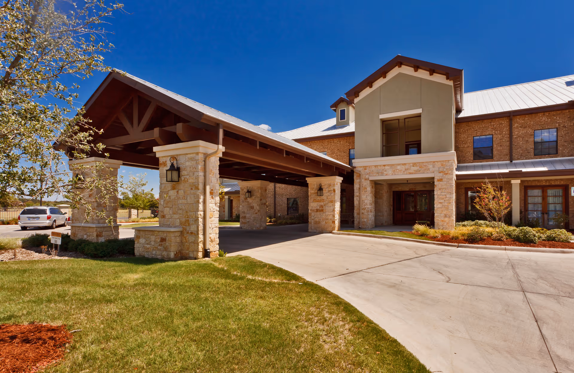 Front entrance of a two-story assisted living building with stone pillars, a covered porte-cochere, and landscaped lawn under a clear blue sky.